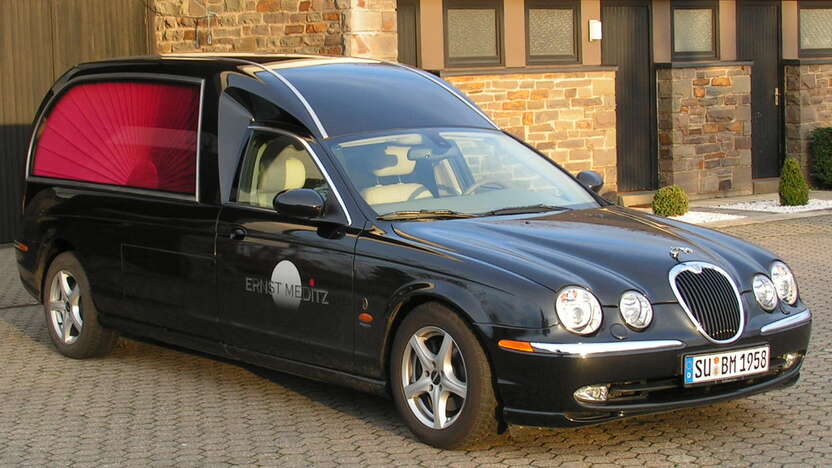 Schwarzer Leichenwagen mit offener Heckklappe, geparkt vor einem modernen Gebäude auf einem gepflasterten Platz.