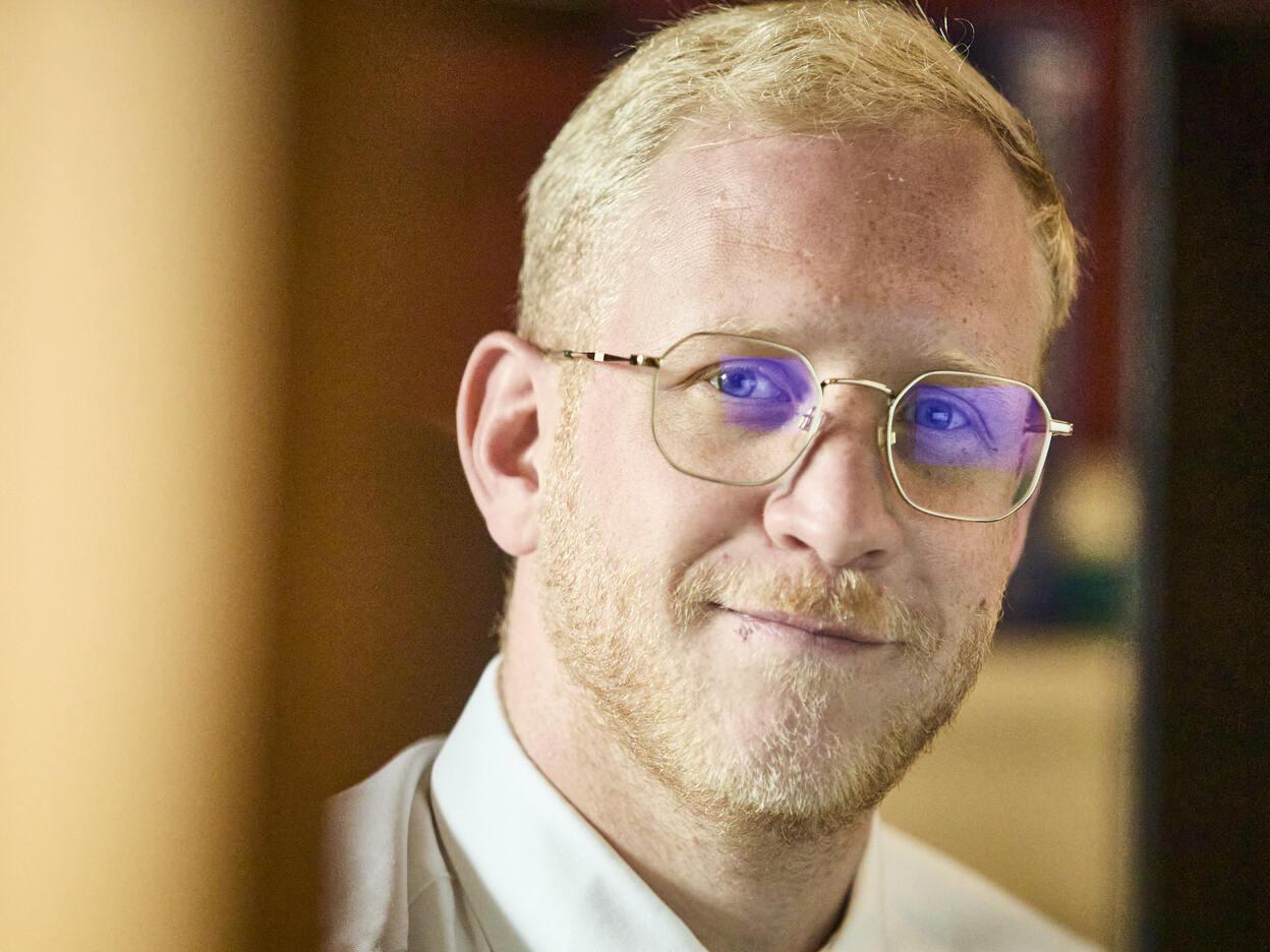 Ein Mann mit Brille und blondem Haar lächelt leicht, während er hinter einem Schreibtisch sitzt.